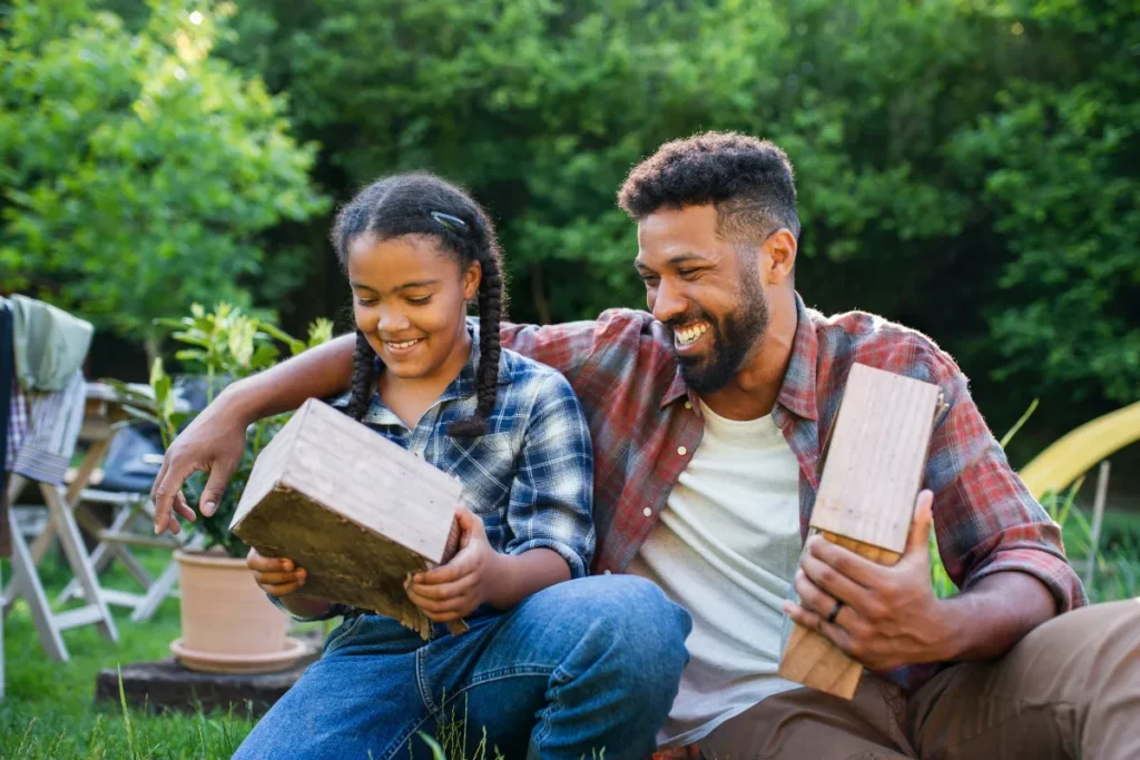 Dad and daughter sit on the grass outdoors, smiling as they hold wooden boxes and work on a craft project.