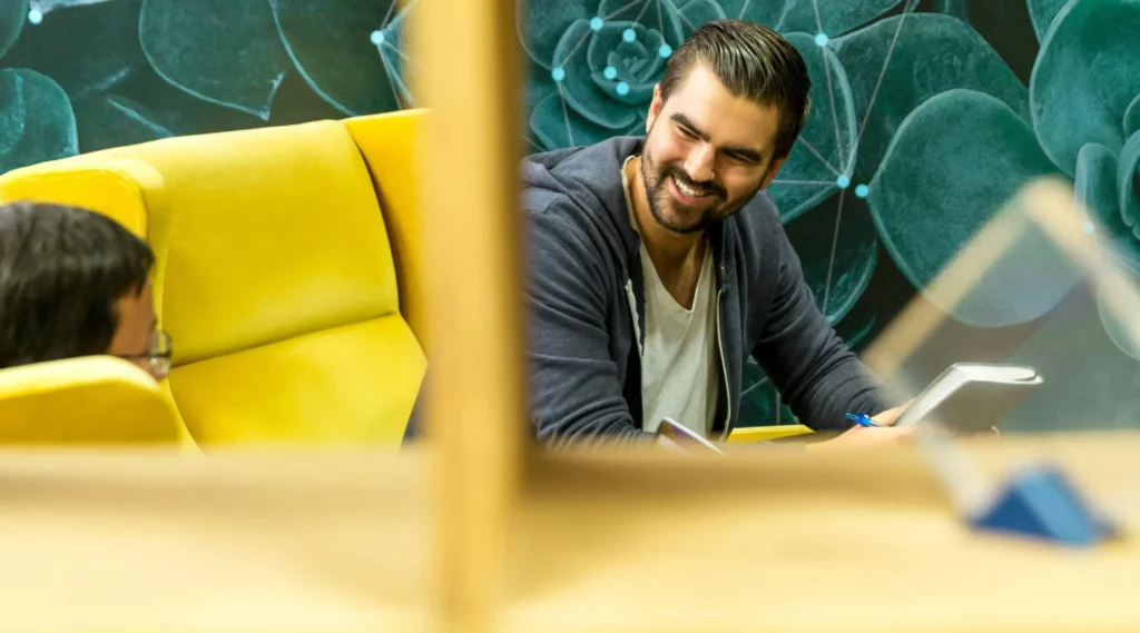 Man with dark hair and beard smiling while writing on a tablet at a wooden table, yellow seating in a green plant-themed background.