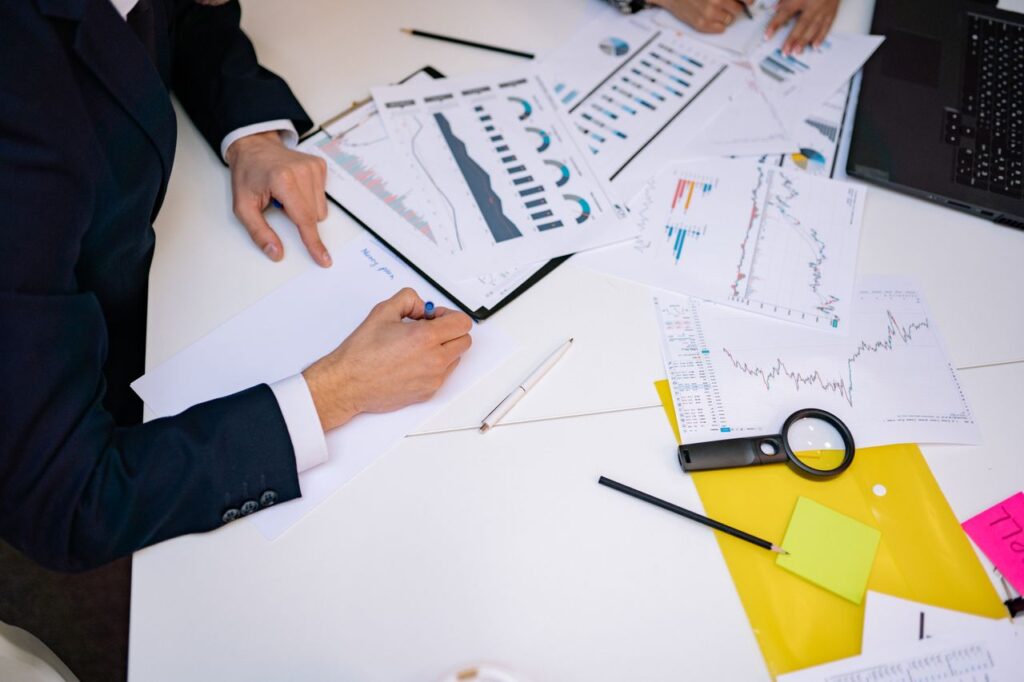 Kierten_Kooner_Finance - Kierten Kooner Hands write on a document amid scattered financial charts, a laptop, and colorful sticky notes on a white table.