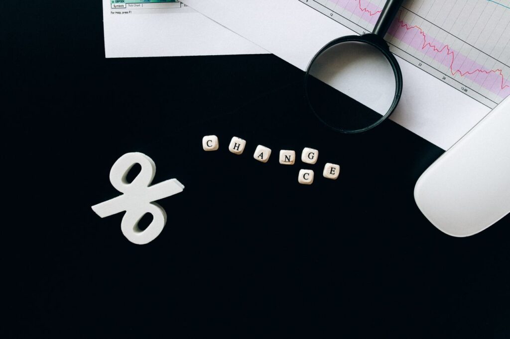Kierten_Kooner_Rates - Kierten Kooner White ampersand icon beside letter cubes spelling CHANGE on a dark desk, with a magnifying glass and chart papers nearby.