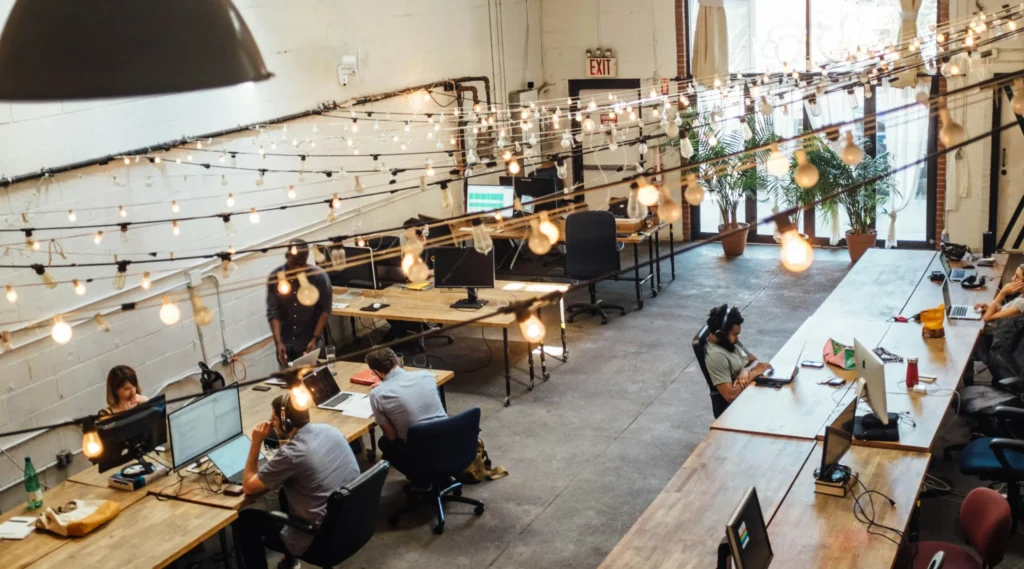 Open-plan coworking space with workers at long wooden desks, laptops, string lights overhead, and potted plants near large windows.