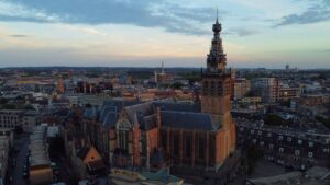 Kierten_Kooner_Dutch_Stylejpg - Kierten Kooner Aerial view of a European city with a tall ornate church tower in the foreground at dusk, skyline in soft light.