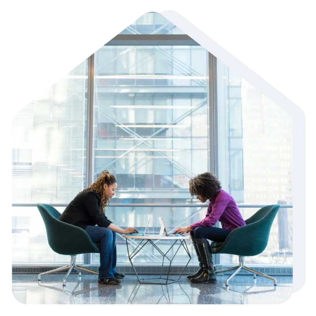 Two coworkers sit across from each other at a glass coffee table, working on laptops in a bright, glass-walled office space.