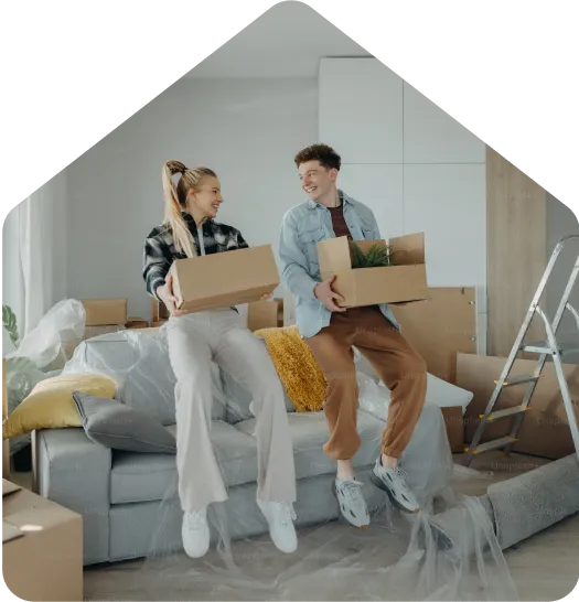 Two people sit on a sofa in a bright living room, smiling while holding moving boxes during a home move, with packing materials and a ladder nearby.