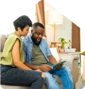 Couple sits on a sofa looking at a tablet together in a cozy living room with a lamp and plants nearby.