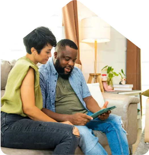 Couple sits on a sofa looking at a tablet together in a cozy living room with a lamp and plants nearby.
