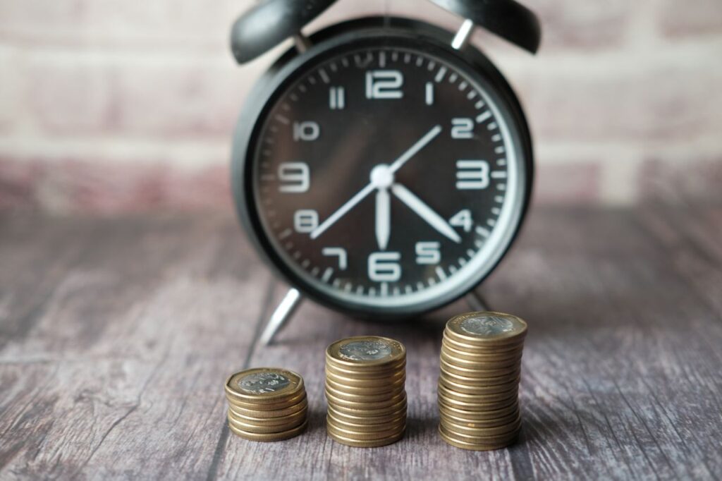 Alarm clock in the background with three stacks of coins in the foreground on a wooden surface, symbolizing time and money (time is money).