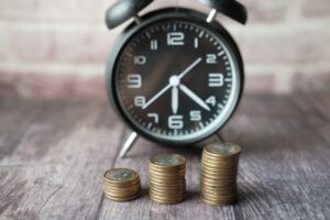 Alarm clock in the background with three stacks of coins in the foreground on a wooden surface, symbolizing time and money (time is money).