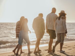 Five friends walk along a sandy beach at sunset, the ocean to their left and warm light behind them.
