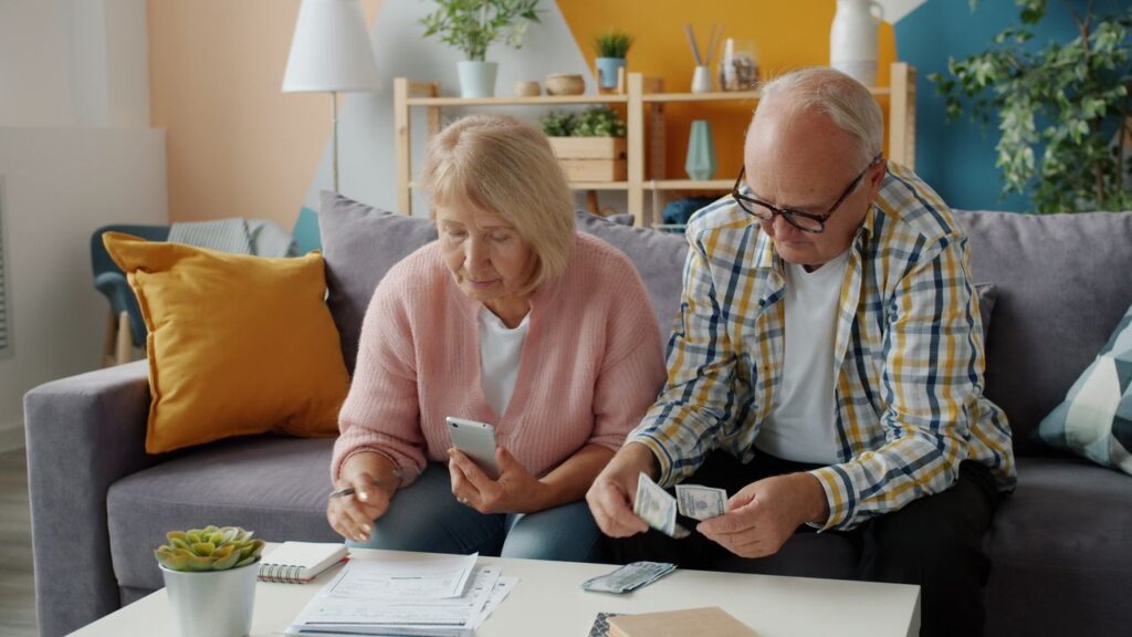 Elderly couple on a gray sofa counting cash and checking documents at a coffee table.