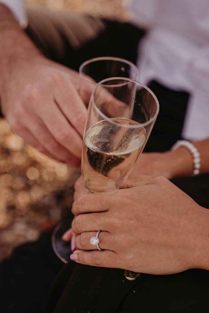 Two people clinking champagne flutes, hands visible with rings and bracelets in a celebratory toast.
